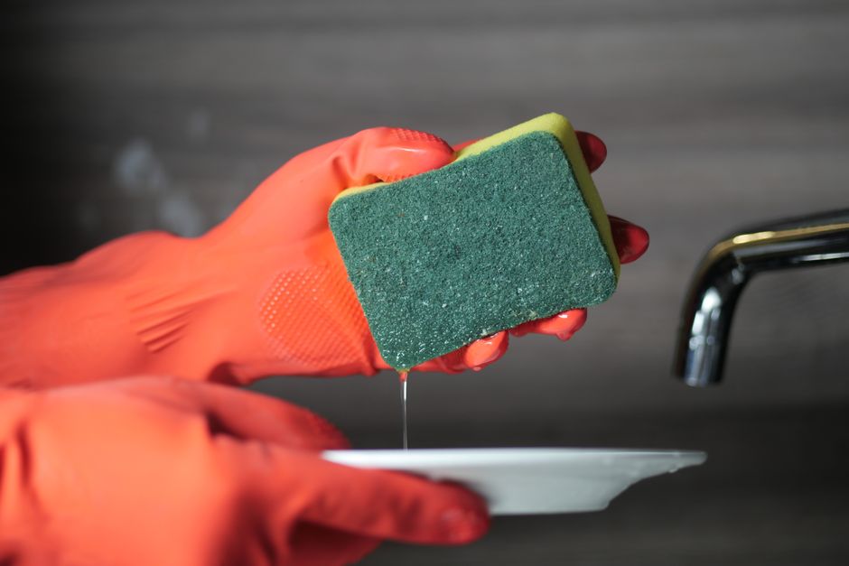Close-up of hands in rubber gloves using a sponge to wash a ceramic plate over a sink.